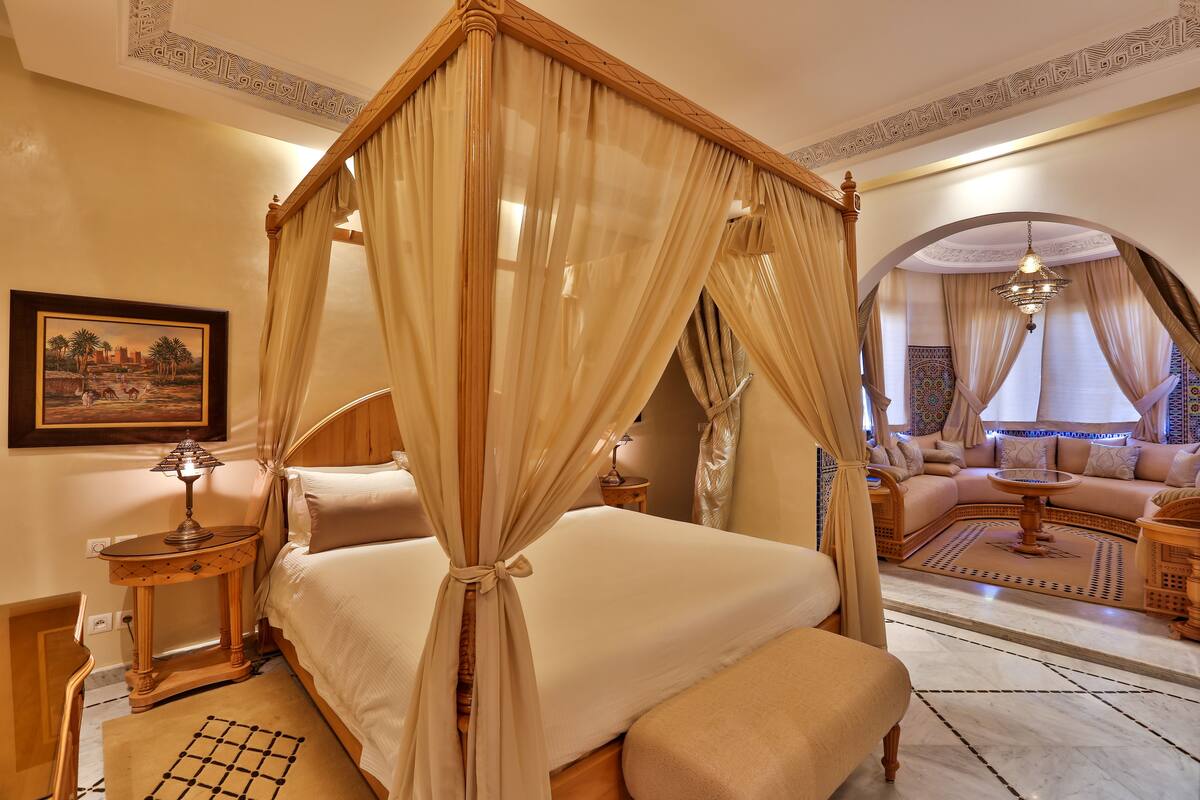 A four-poster bed with sheer curtains, a bedside table with a lamp, a framed picture on the wall, and a sofa set in the background.