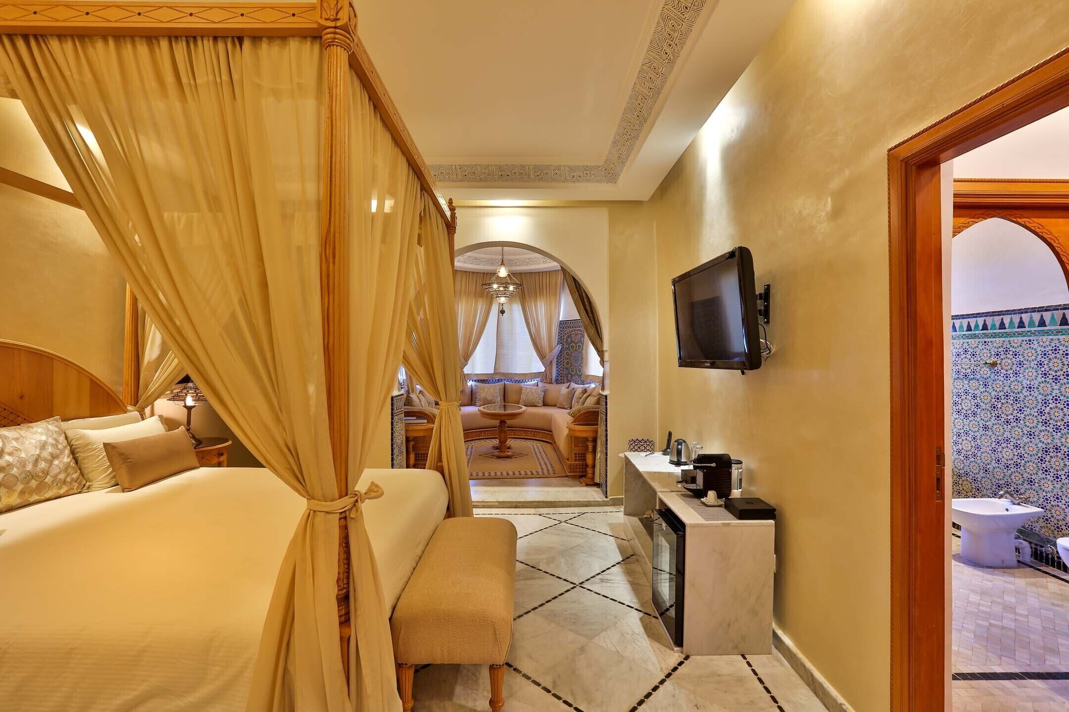 A hotel room with a bed, a bench, a TV, a small table, and a bathroom visible through an archway.