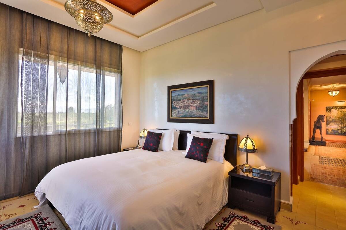 A large bed with white linens and two decorative pillows, a nightstand with a lamp, a framed painting on the wall, and a view of the outdoors through a window with sheer curtains.