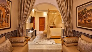 Habitación de hotel con cama, zona de estar con sofá, escritorio y televisión. Hay cortinas y cuadros en las paredes.