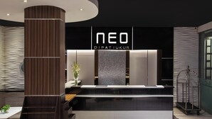 Un área de recepción moderna con un llamativo letrero 'NEO DIPAT IUKUR', una mesa de recepción, una planta y un carrito metálico.