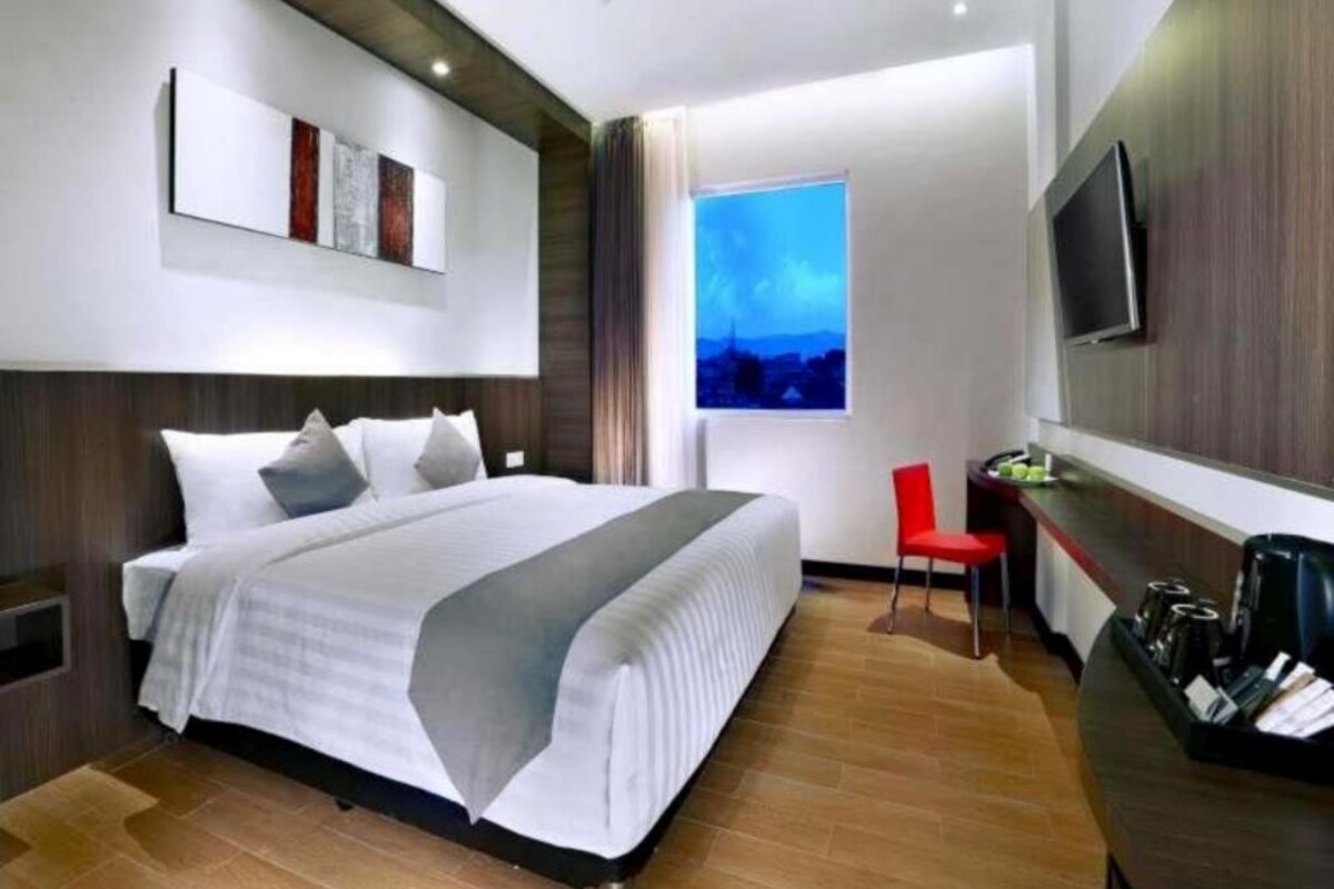 A modern hotel room with a large bed, a desk with a chair, a flat-screen TV, and a window with a cityscape view.