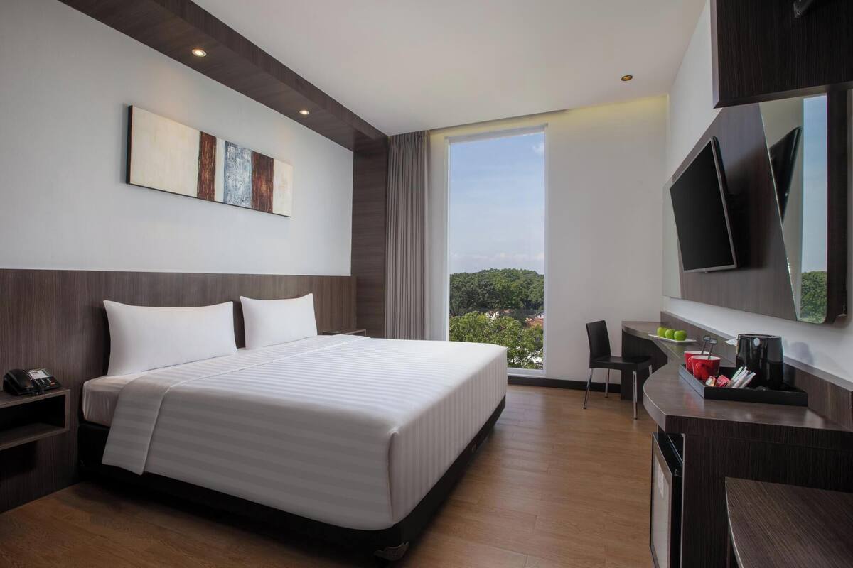 A modern hotel room with a large bed, a desk, a chair, a flat-screen TV, and a large window offering a view of the outdoors.
