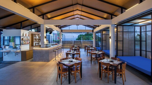 Un amplio comedor interior con varias mesas y sillas, una zona de bar con estanterías llenas de botellas y grandes ventanales con vistas al exterior.