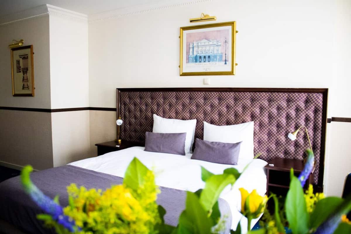 Een hotelkamer met een groot bed, een hoofdbord met een geruit patroon, een ingelijste foto aan de muur en een nachtkastje met een lamp.