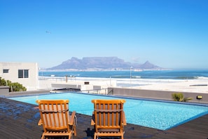 A pool with two wooden chairs facing a beach and mountains.