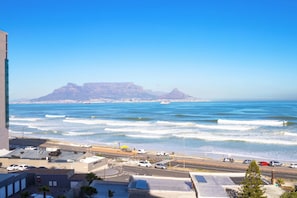 A coastal cityscape with a prominent mountain in the background, a busy street with cars, and a clear blue sky.