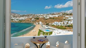 Un balcón con vistas a una playa y una piscina, dos sillas blancas y una mesa redonda pequeña con una copa de vino.