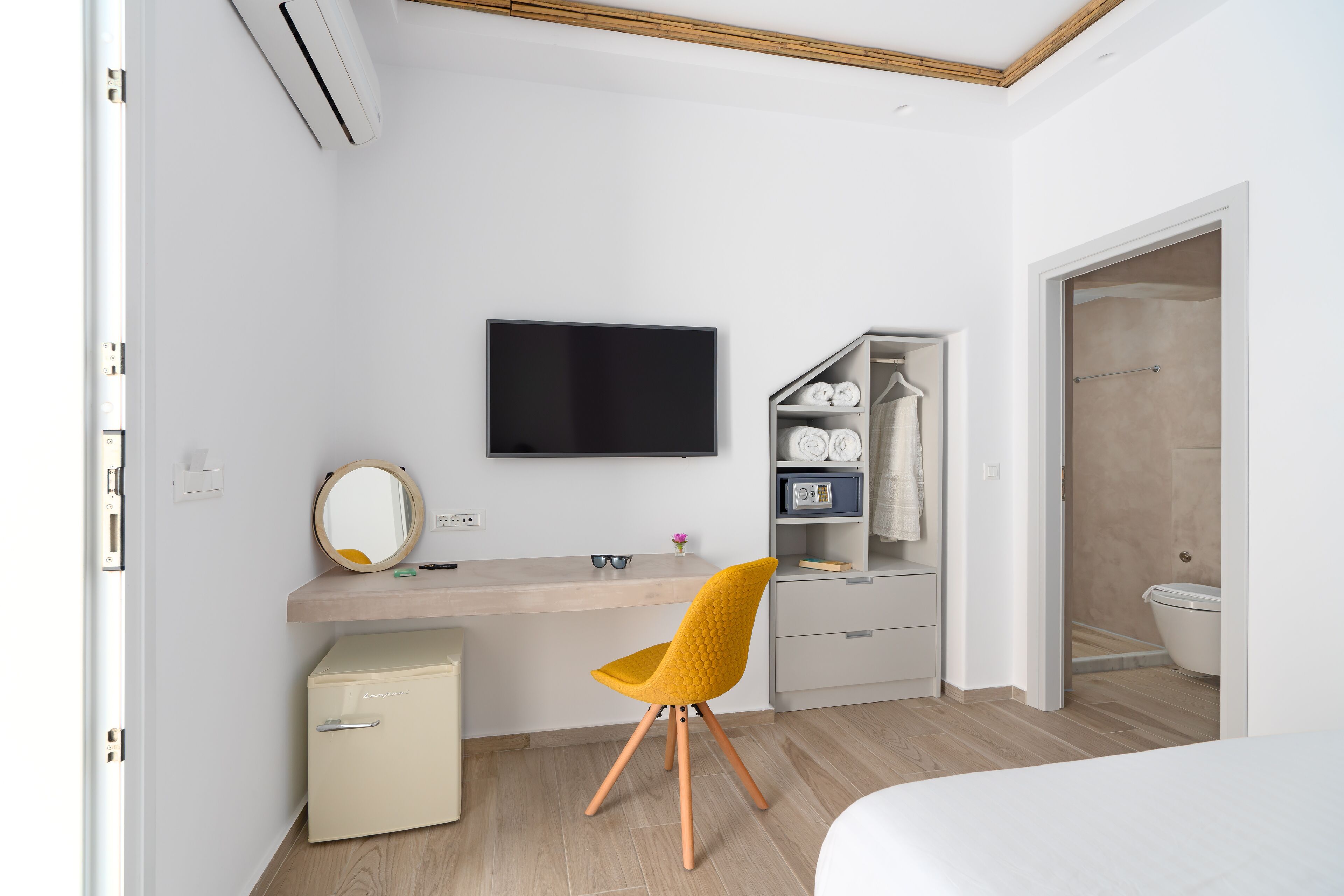 A modern bedroom with a bed, a desk with a computer, a chair, a TV, a wardrobe, and a small fridge.