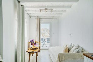 A room with a bed, a chair, a table, and a view of the outside. - Cape Mykonos (Mykonos)