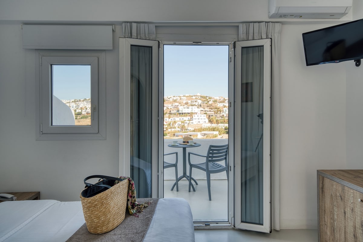 A hotel room with a bed, a small table with chairs, a TV, and a view of a hillside town.