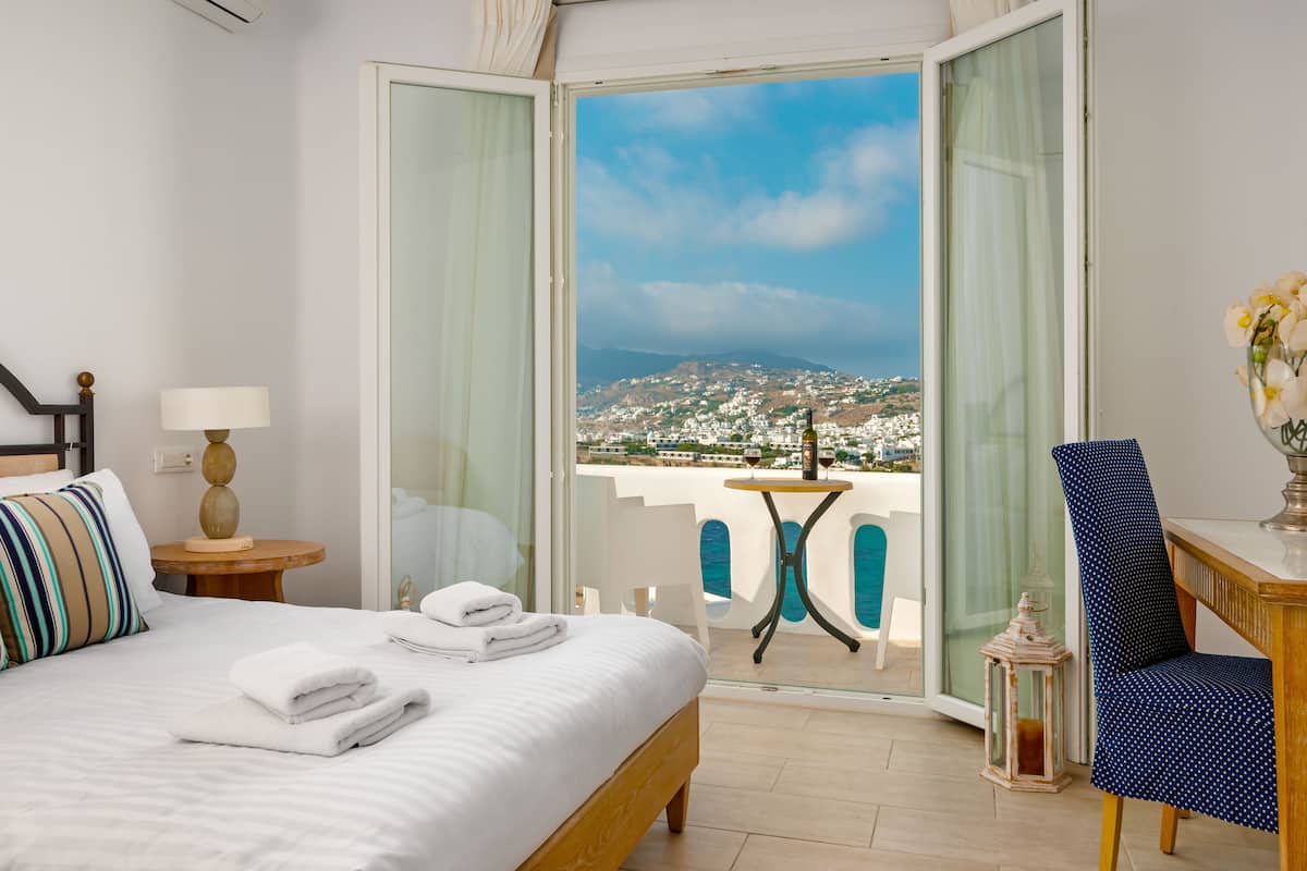 A hotel room with a bed, a table with two wine glasses, a chair, and a view of a coastal town.