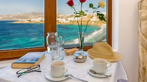 Una mesa con un libro, gafas, dos tazas, un florero con rosas, un sombrero y vista al mar a través de una ventana.