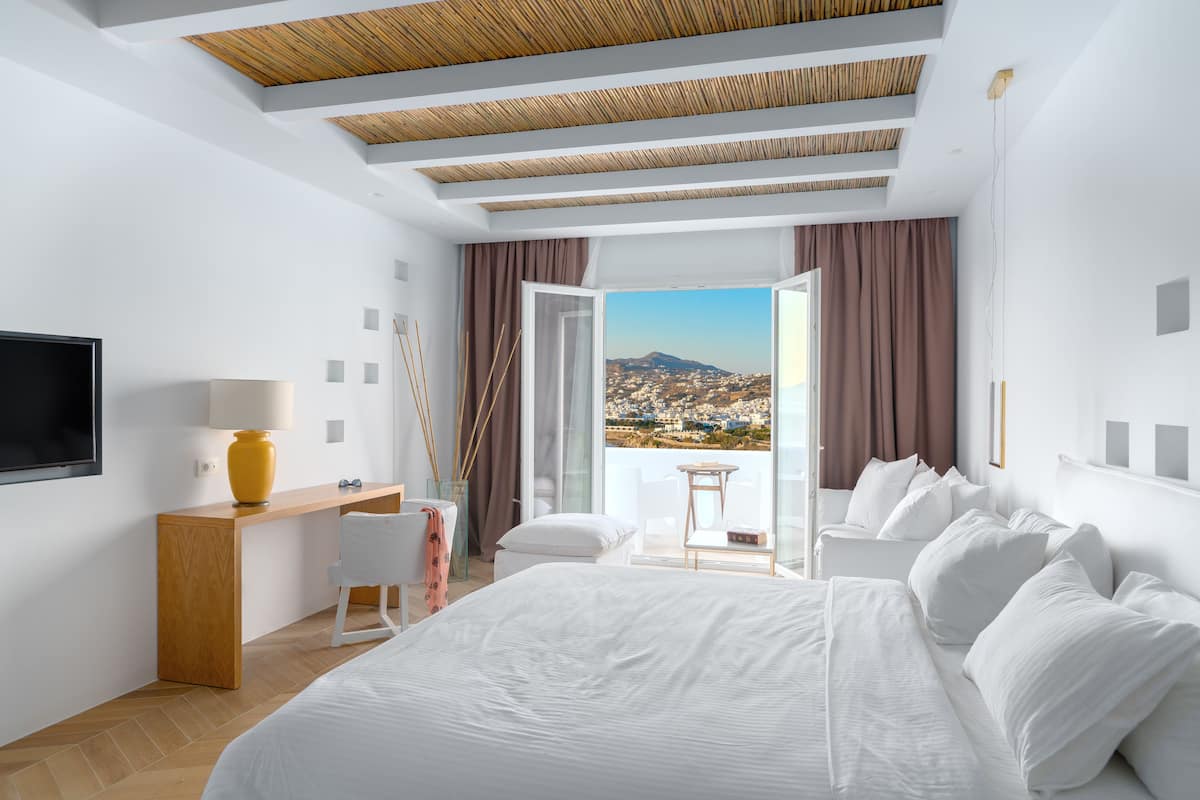 A modern hotel room with a large bed, a desk, and a view of a cityscape through a balcony.
