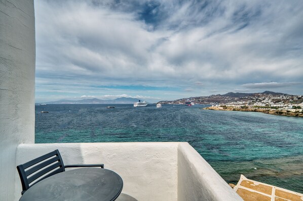 A balcony with a table and chairs overlooking a coastal area with ships and a town. - Cape Mykonos (Mykonos)
