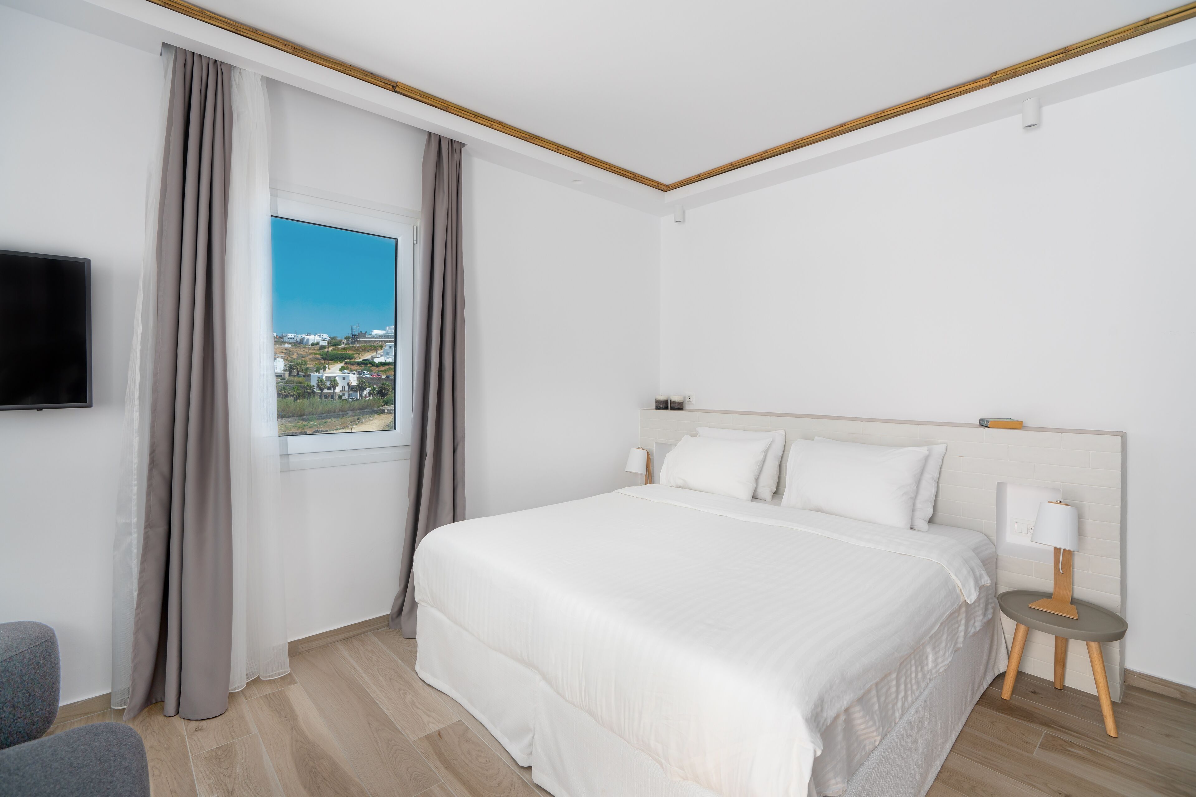 A hotel room with a large bed, a TV mounted on the wall, a window with curtains, and a bedside table with a lamp.