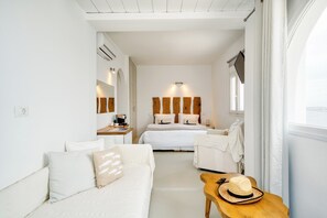 A hotel room with a bed, a sofa, a small table with a hat, and a window. - Cape Mykonos (Mykonos)