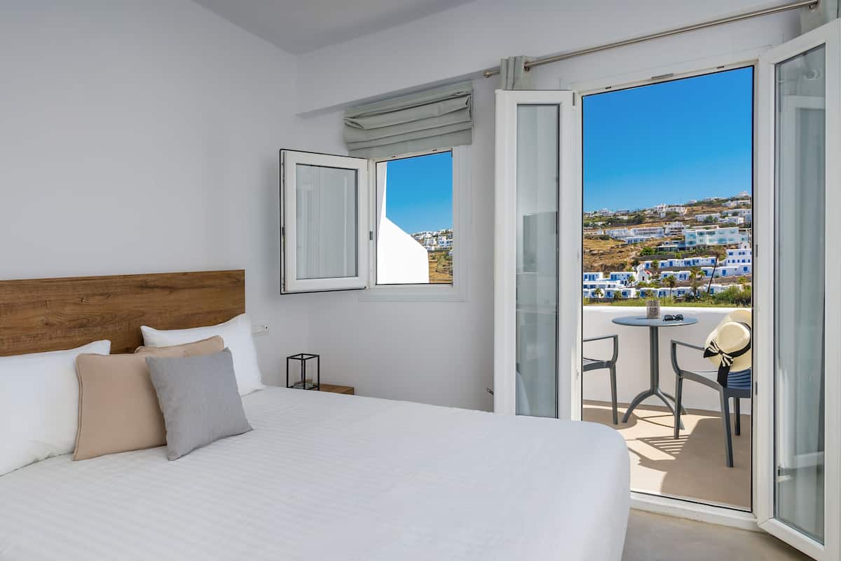 A bedroom with a large bed, a window with curtains, a view of a hillside, and a balcony with a table and chairs.