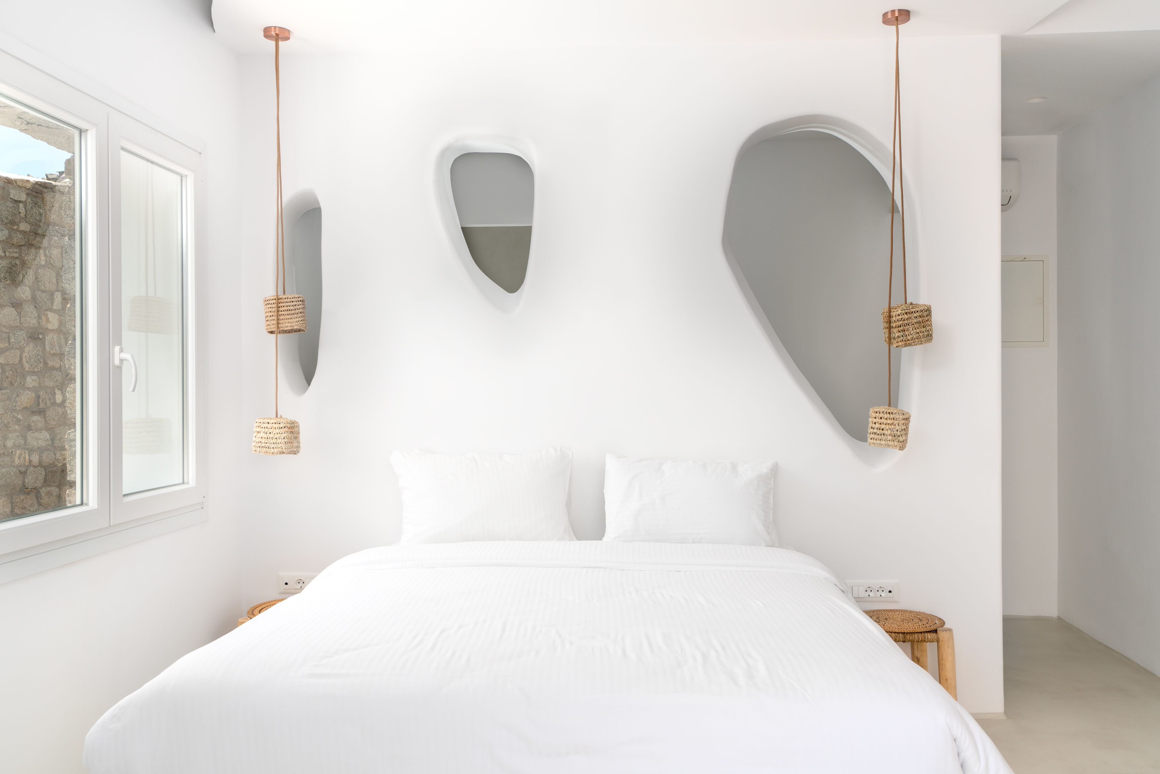 Une chambre à coucher minimaliste avec un grand lit, du linge de lit blanc et un décor mural original composé de miroirs et d’appliques lumineuses tressées.