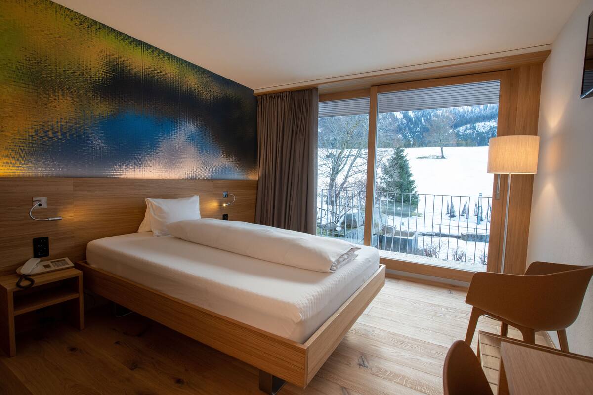 Een hotelkamer met een houten bed, een nachtkastje, een stoel en een groot raam met uitzicht op een besneeerd landschap.