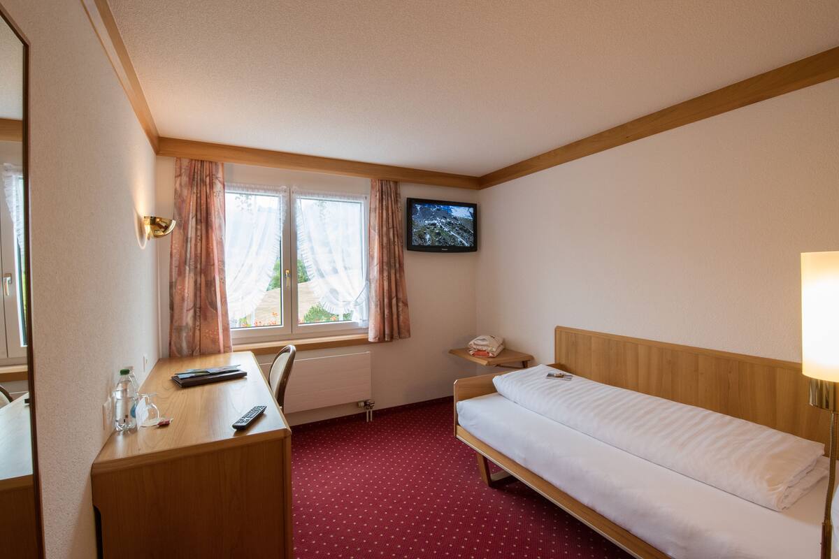 Een hotelkamer met een bed, een bureau met stoel, een aan de muur bevestigde televisie en een raam met gordijnen.