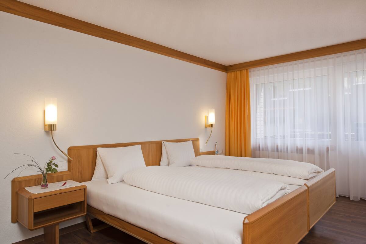 Hotelkamer met een houten bed, twee nachtkastjes met lampen, een bijzettafeltje en een raam met dunne gordijnen.