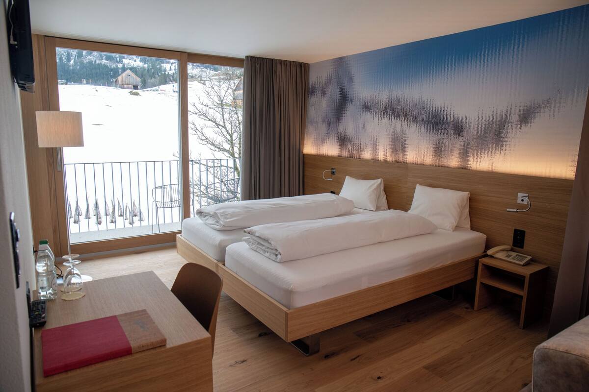 Een hotelkamer met een groot bed, een bureau met een stoel, een klein tafeltje, en uitzicht op een besneeerd landschap door een groot raam.