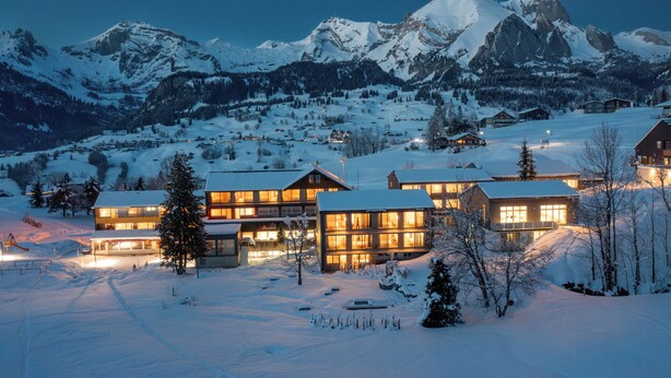 A snow-covered mountain resort with multiple buildings, ski slopes, and a clear view of the mountains.