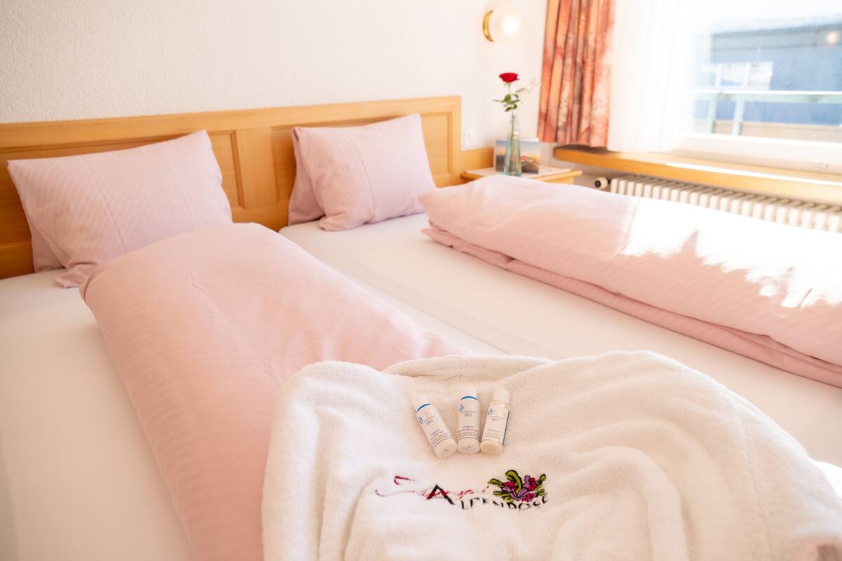 Hotelkamer met twee bedden, elk met roze beddengoed en een houten hoofdbord. Er ligt een witte badjas met geborduurd tekst op het bed, samen met drie flessen.