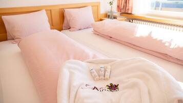 A hotel room with two beds, each with pink bedding and a wooden headboard. There is a white bathrobe with embroidered text on it, and three bottles on the bed.