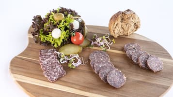 Ein Holzbrett mit verschiedenen Lebensmitteln, darunter geschnittene Salami, Gurken, ein Brotlaib, ein Salat mit Salat, Tomaten und Oliven.