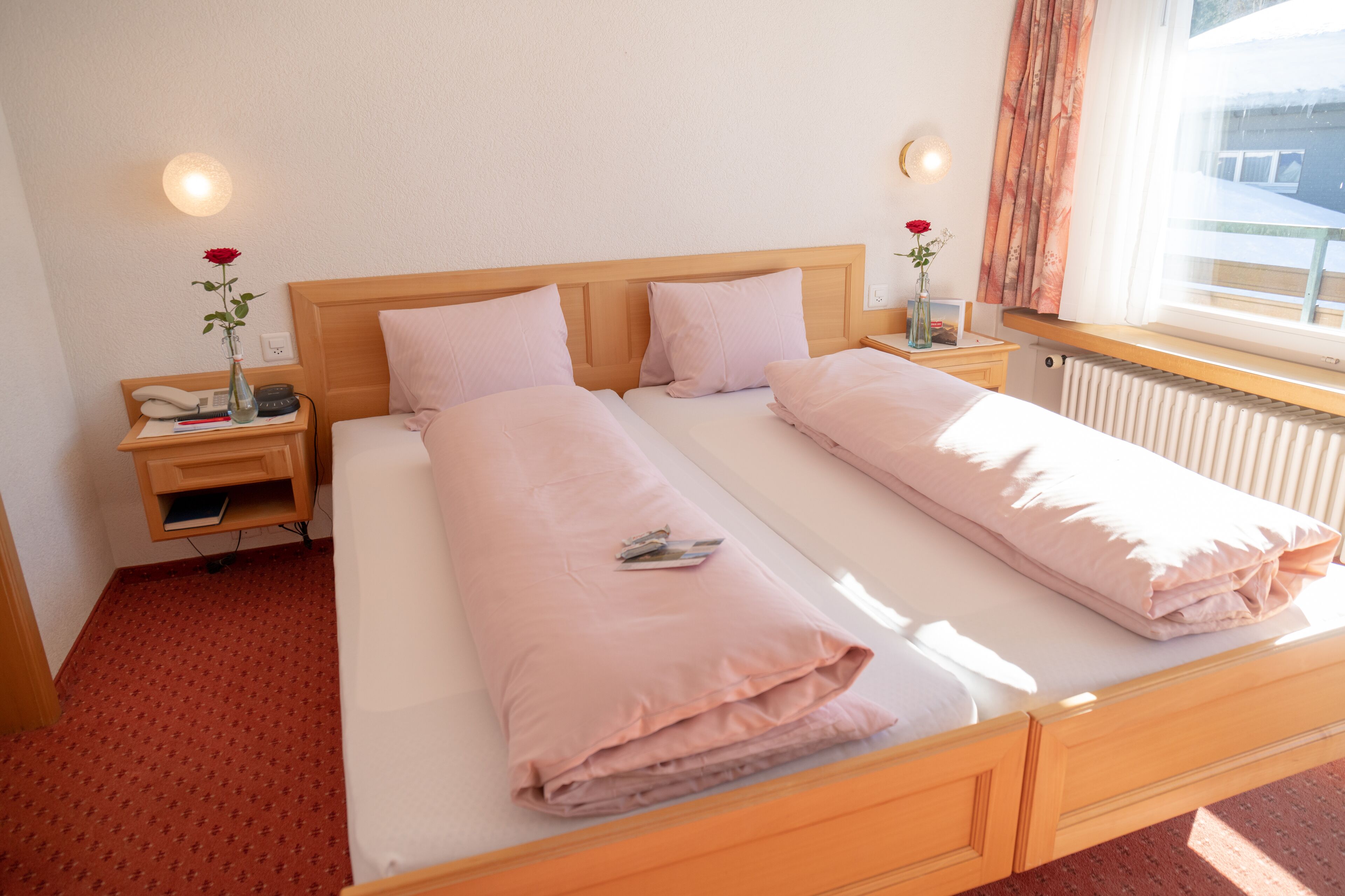A double bed with a wooden headboard, two bedside tables with lamps, a vase with a red rose, and a window with curtains.