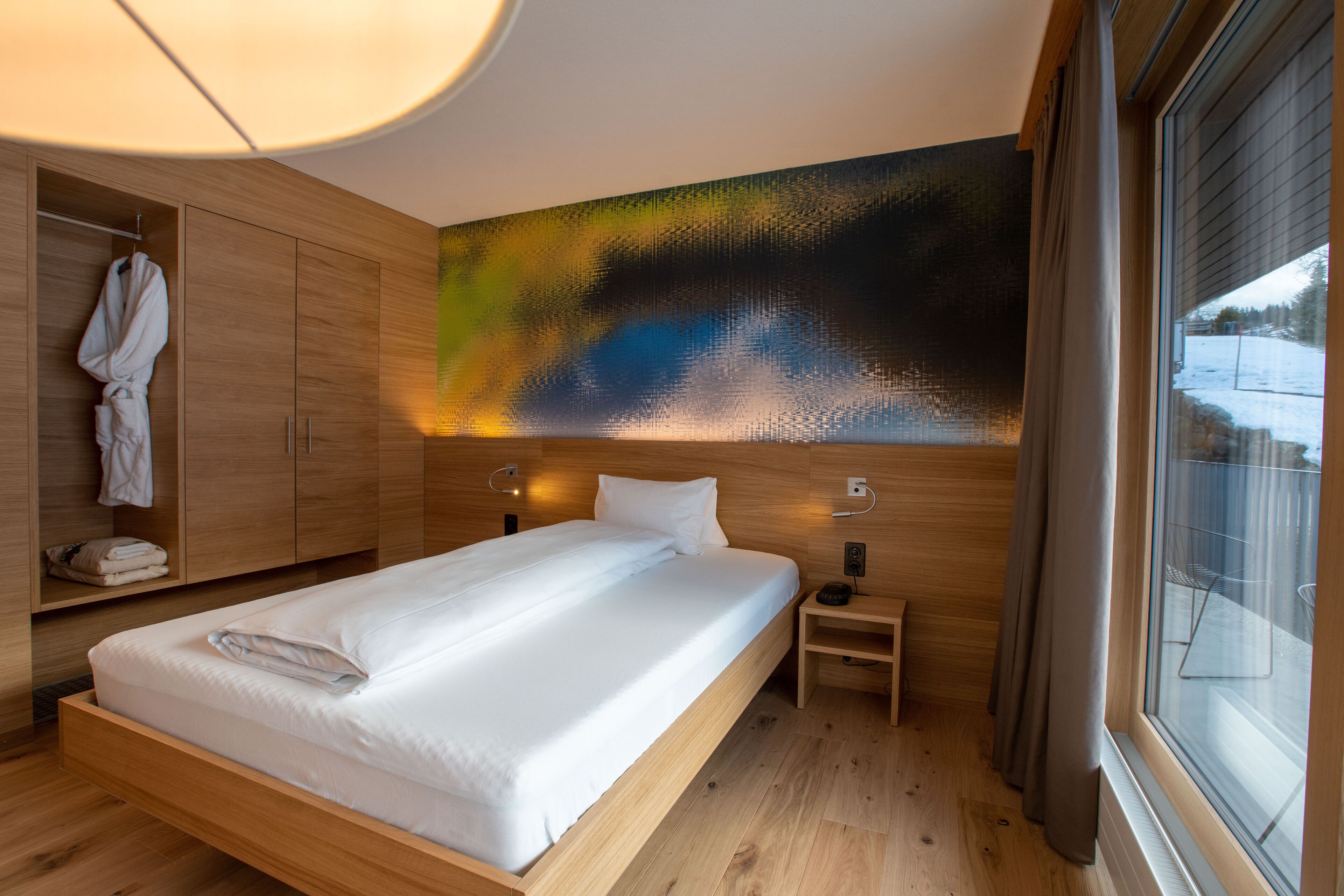 Une chambre d’hôtel moderne avec un lit en bois, un grand tableau abstrait accroché au mur et une vue sur un paysage enneigé par la fenêtre.