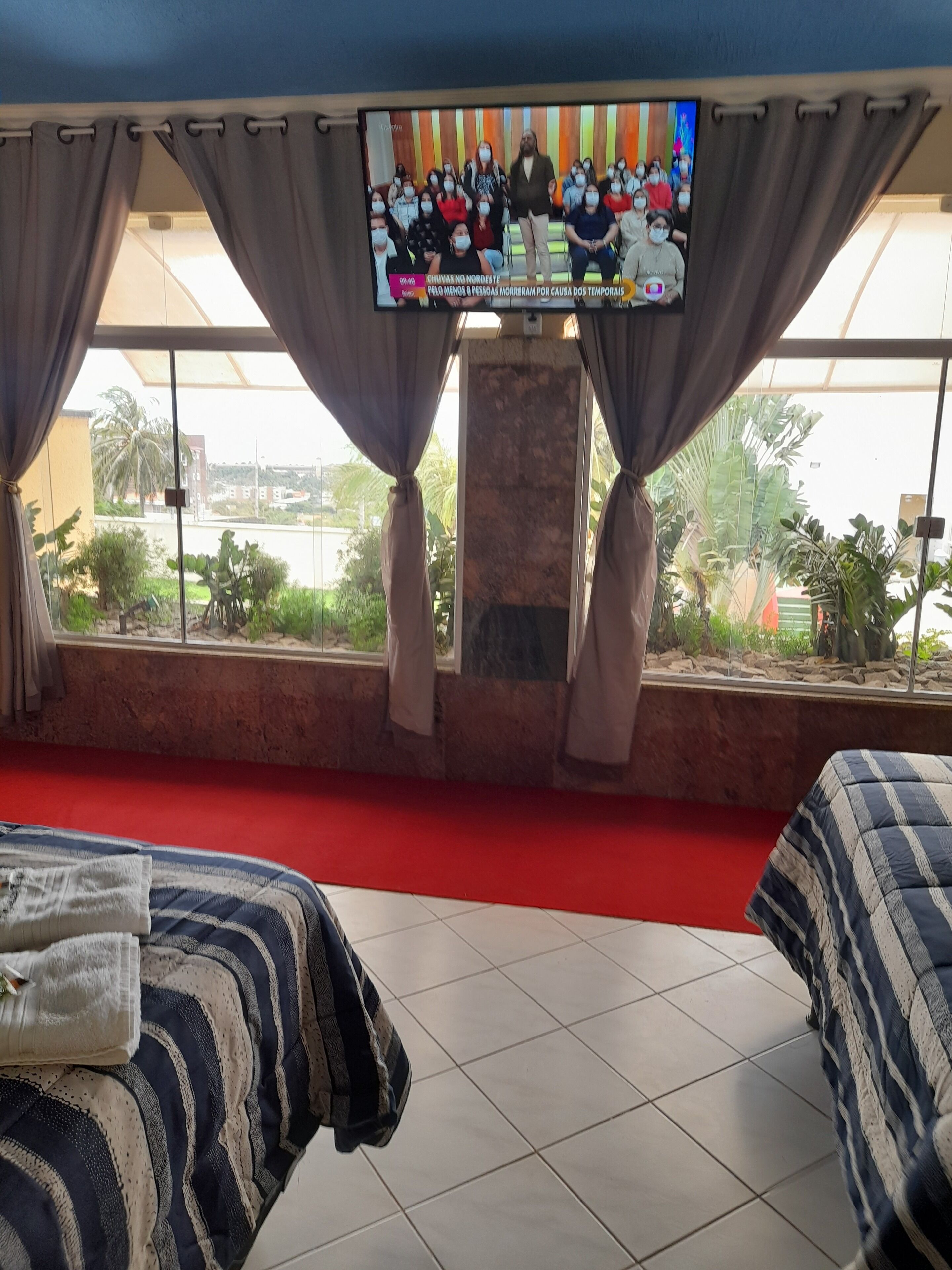 Habitación de hotel con una gran pantalla de televisión mostrando un noticiero, una alfombra roja y vistas al exterior a través de amplias ventanas.