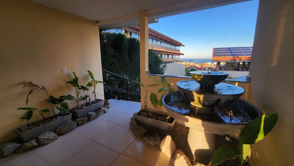 A balcony with potted plants, a tiered water fountain, and a view of a building with solar panels. - Pousada Ceará Porto das Dunas (Aquiraz)