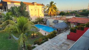 A swimming pool surrounded by palm trees and a thatched-roof structure, with a view of distant buildings and power lines. - Pousada Ceará Porto das Dunas (Aquiraz)