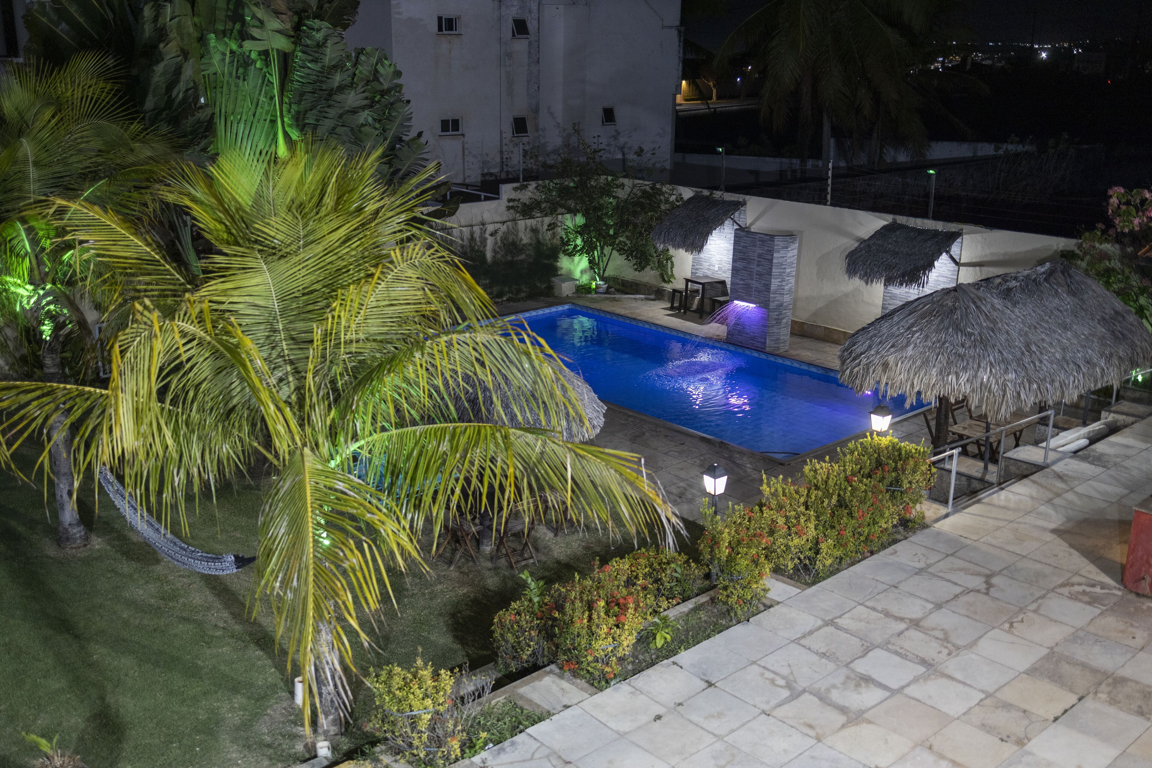 A swimming pool with a thatched-roof structure, surrounded by palm trees and a well-lit patio area.