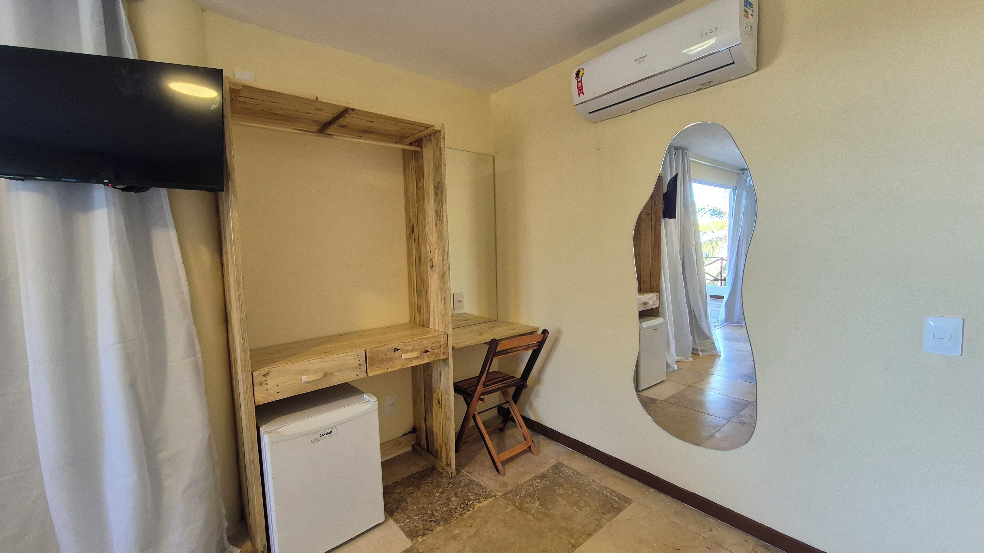 A small, compact room with a wooden desk, a white refrigerator, and a mounted air conditioner.
