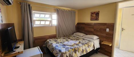 A bedroom with a bed, a desk with a computer, an air conditioner, a window with curtains, and a door.