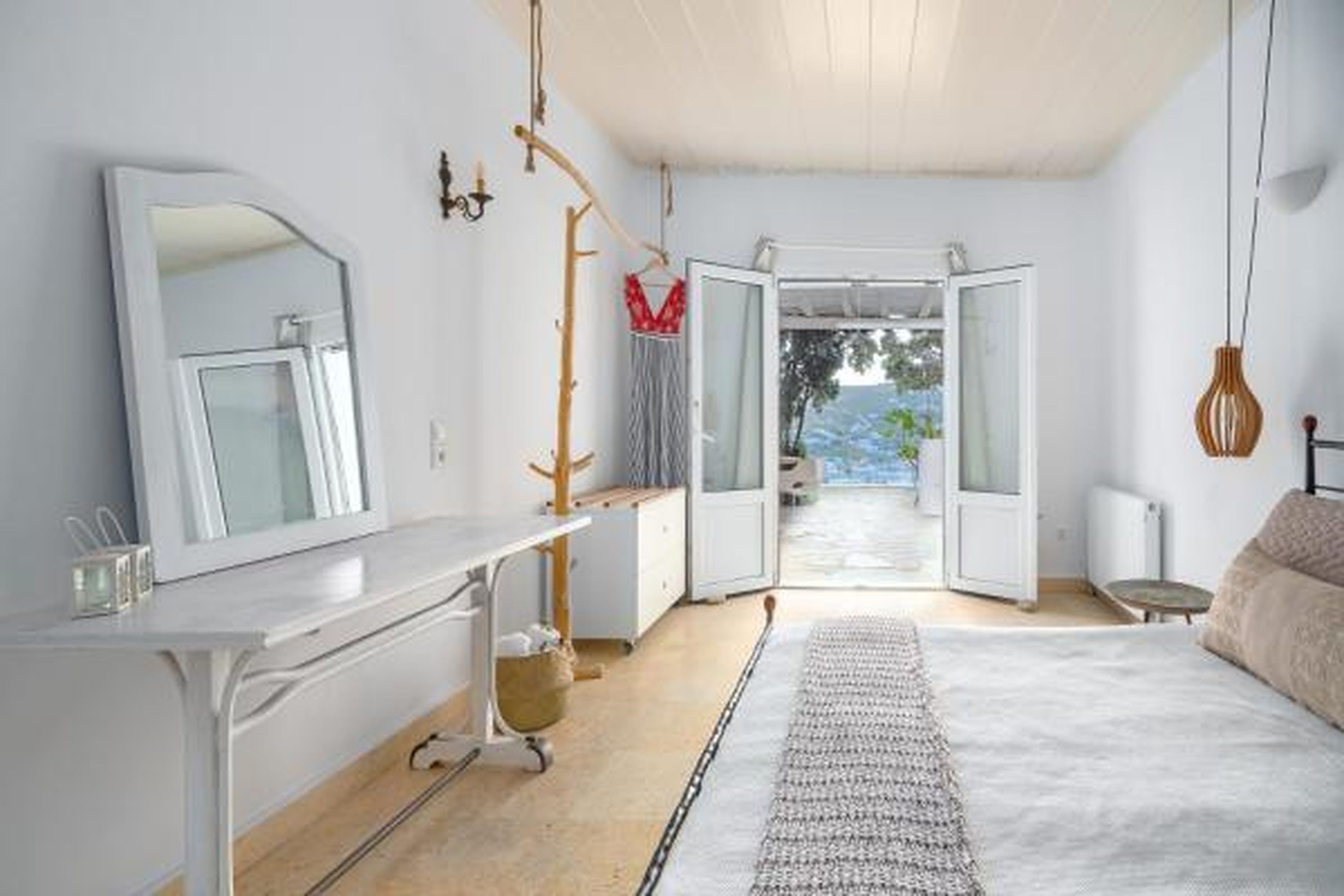 A modern bedroom with a large bed, a bedside table, a mirror, and an open door leading to a balcony with a view of the outdoors.