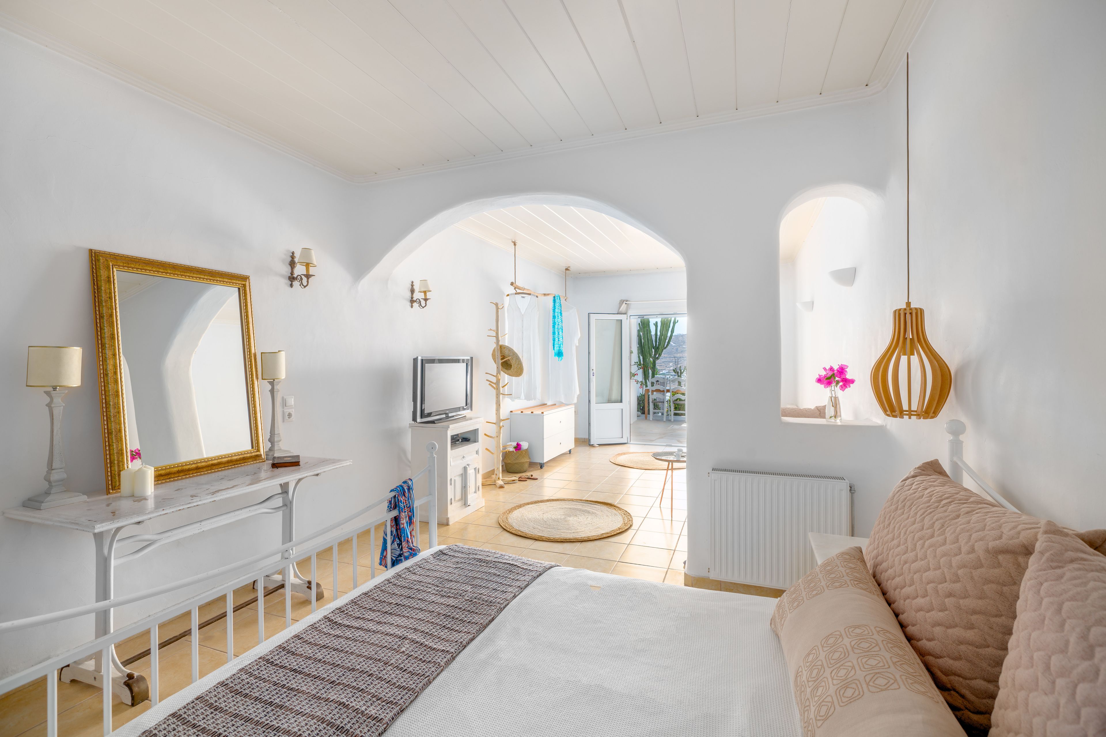 A bright, spacious bedroom with a large bed, a white vanity with a mirror, and a view of the living area through an archway.
