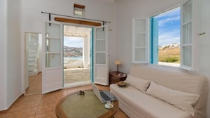 A room with a white sofa, a laptop on a small table, and a view of a coastal landscape through open doors. - Ornos Blue (Mykonos)