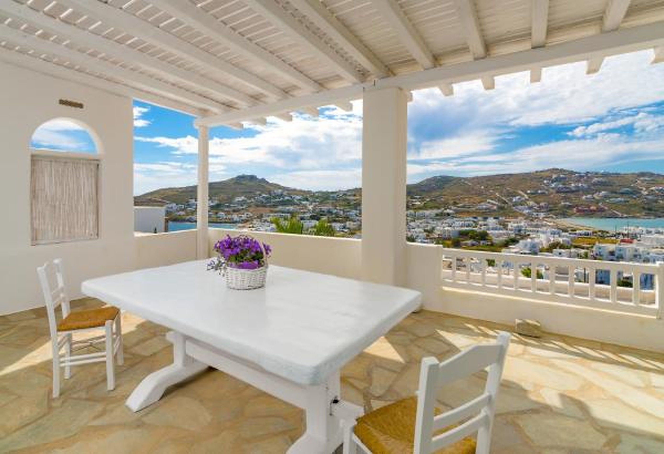 A terrace with a white table and chairs, a basket of flowers, and a view of a coastal town.