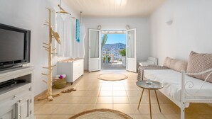 A room with a TV, a wooden drying rack, a bed, a small table, and a view of the city through an open door. - Ornos Blue (Mykonos)