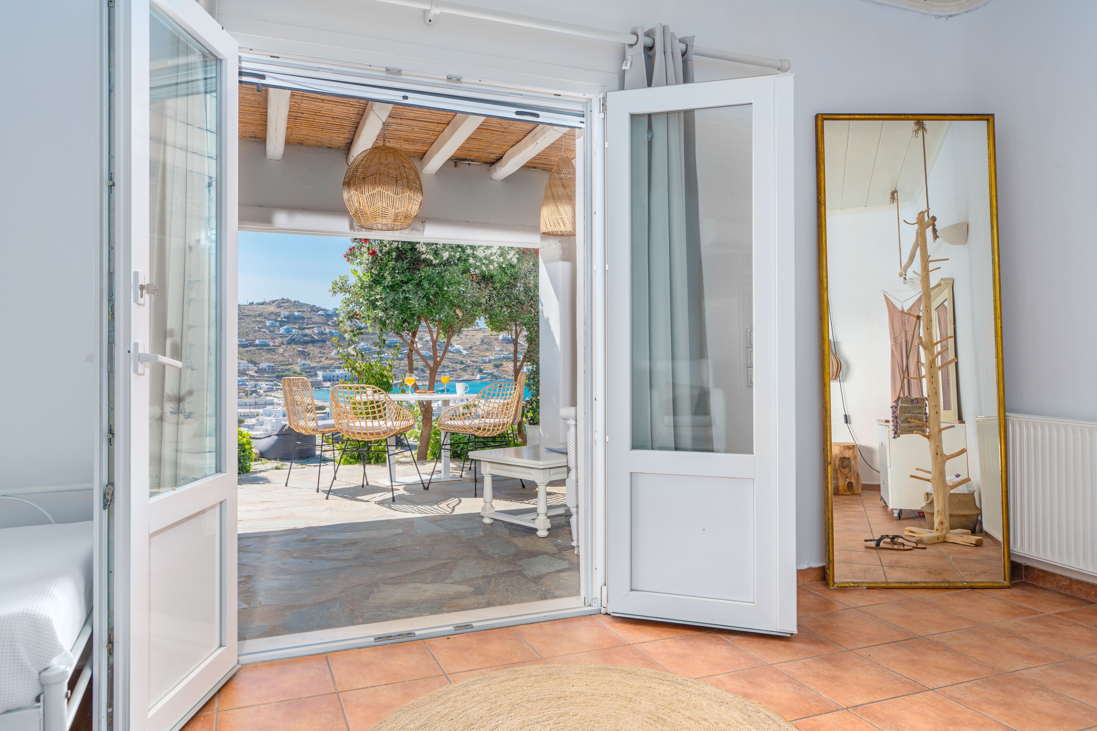 An open sliding glass door leading to an outdoor patio with a table and chairs, a view of a hillside, and a large mirror on the wall.