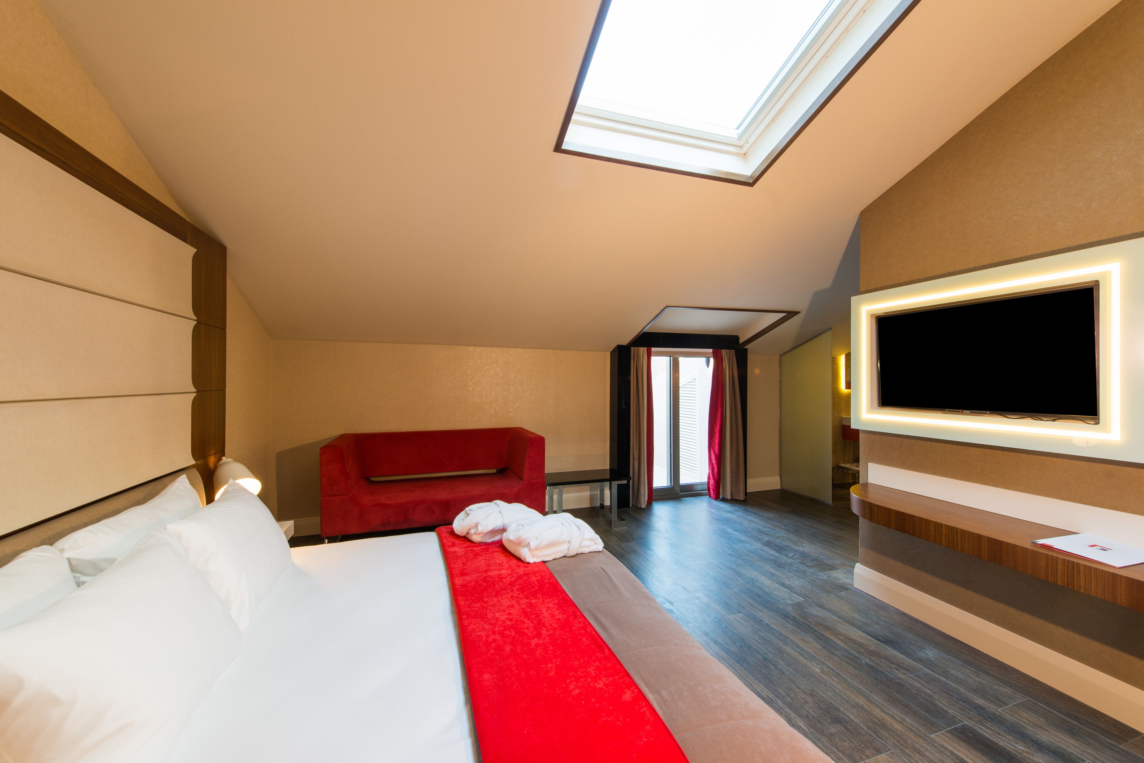 A hotel room with a bed, a red sofa, a TV, and a skylight.