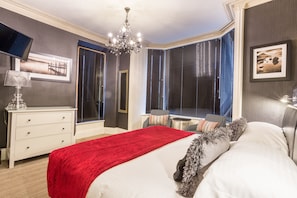 Une chambre à coucher avec un grand lit, un plaid rouge, un lustre, une télévision et une fenêtre avec des stores.