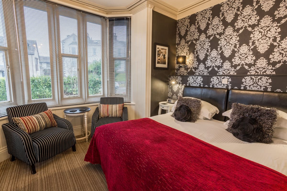 A hotel room with a large bed, a red throw blanket, striped armchairs, a small round table, and a wall with a floral pattern.