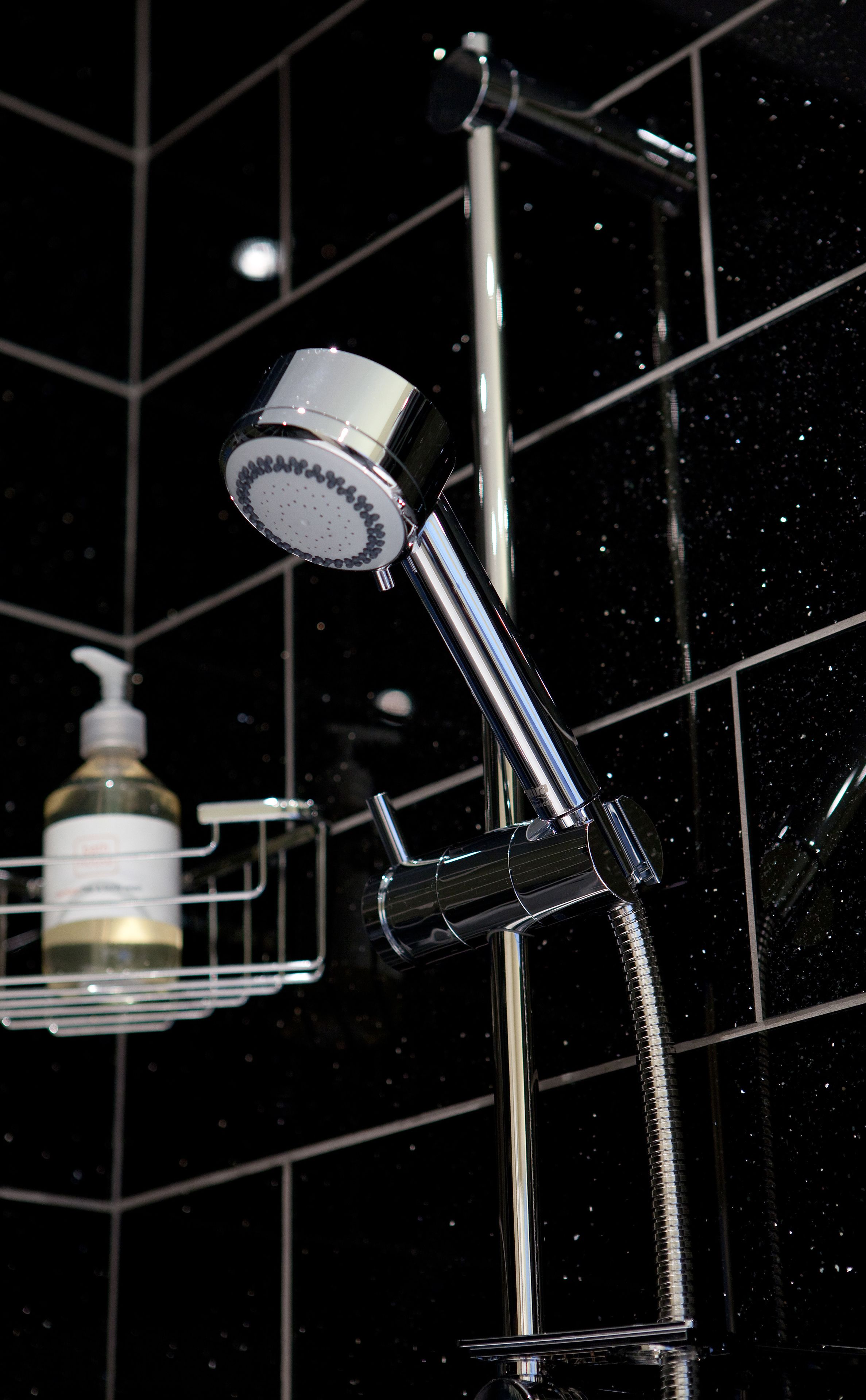 A modern bathroom with a chrome shower head and a handheld shower attachment. A soap dispenser is visible on a small shelf.