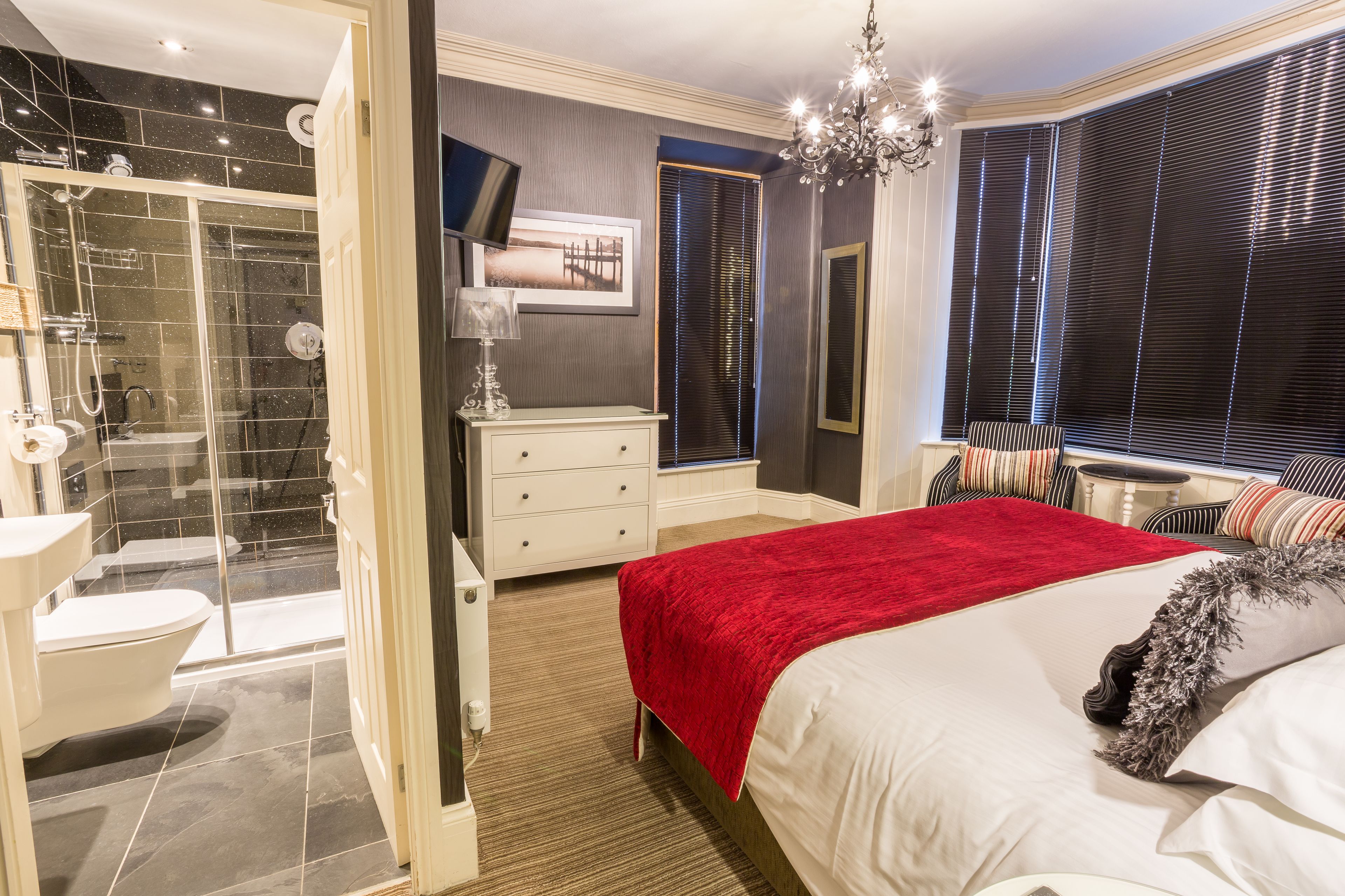 A modern bedroom with a large bed, a red throw blanket, a chandelier, a vanity, and a shower area.